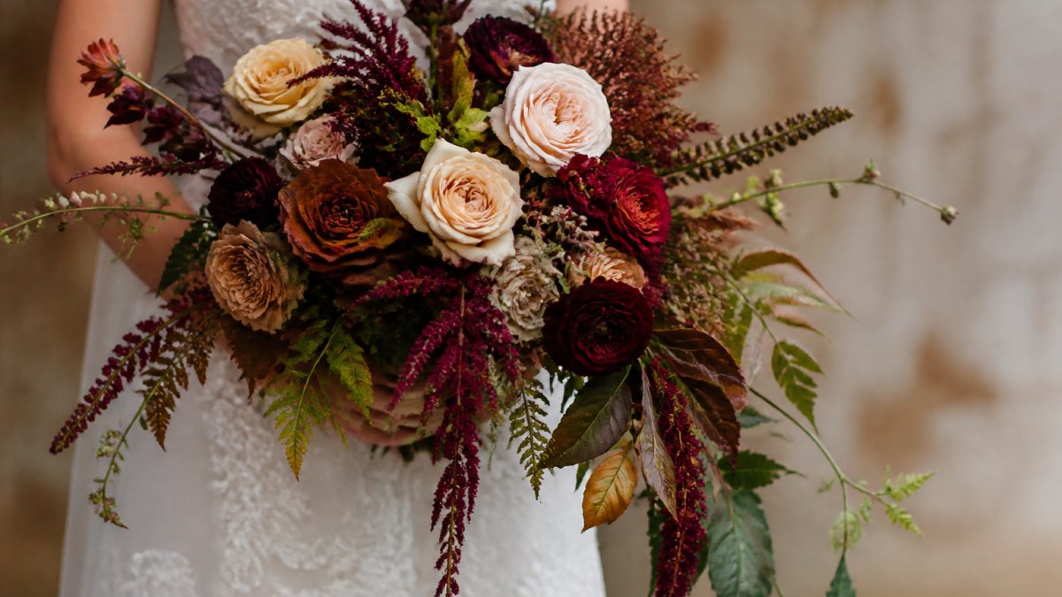 Close-up of a sculptural bridal bouquet featuring garden roses, burgundy blooms, and trailing amaranthus against a soft neutral background, showcasing rich earth tones and modern arrangement technique