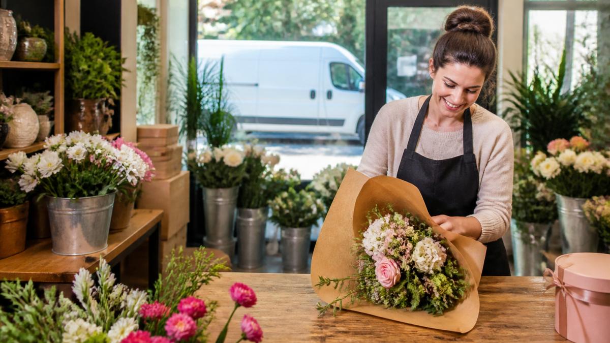A florist in a bright local flower shop wrapping a fresh bouquet in kraft paper, with buckets of seasonal stems and a delivery van visible through the front window