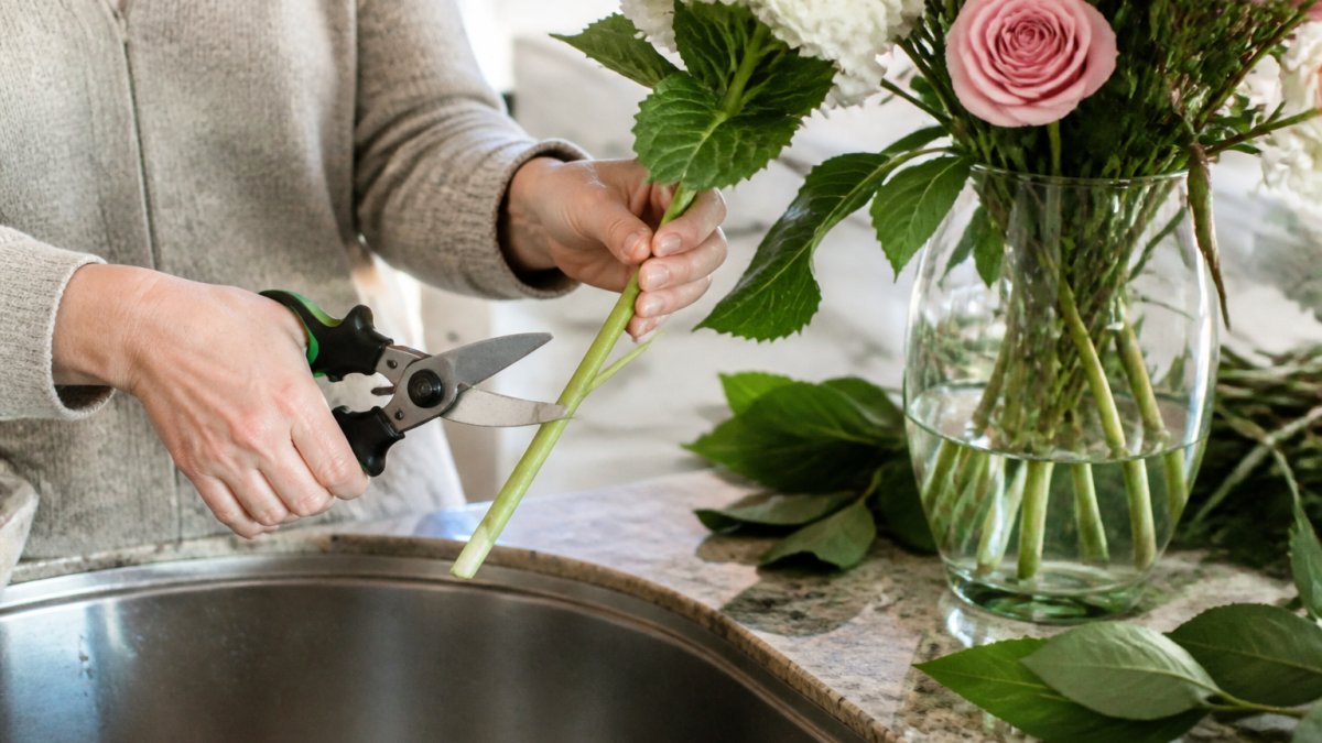 Close-up of hands holding floral shears cutting a flower stem at a 45-degree angle over a kitchen sink, with a glass vase of fresh flowers and loose green leaves on the counter nearby